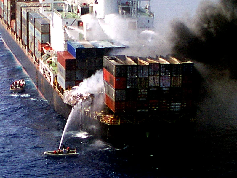 Navio porta-contêineres em chamas no mar, com barcos jogando água